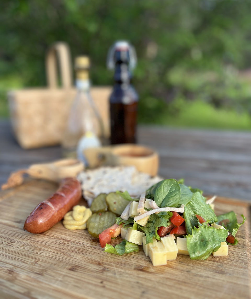 A board lying on a table outside with salad, small pieces of cheese, pickled cucumber slices, crispbread, mustard and sausages with two wooden cups, two glass bottles and a basket in the background.