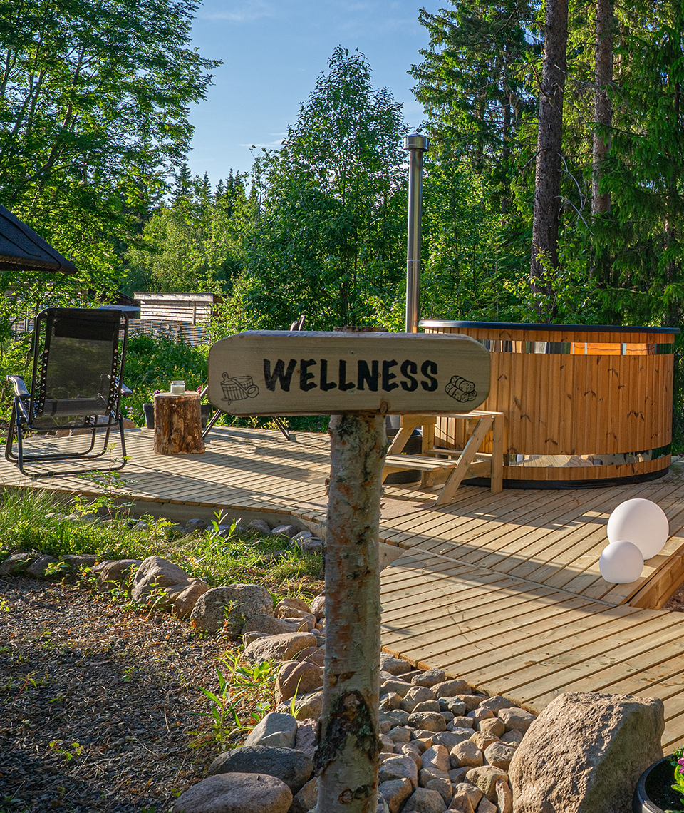 There's a wooden sign that says Wellness with a wooden platform on which there are sun loungers, a few lamps and a hot tup, and green trees in the background.
